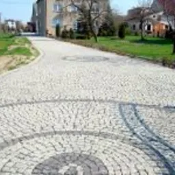 Szeroki podjazd wyłożony jasną kostką brukową z dekoracyjnymi okręgami z ciemniejszej kostki, prowadzący do domu jednorodzinnego w tle.